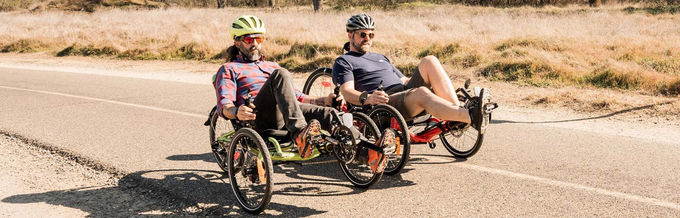 Two men riding recumbent trikes next to each other on a trail