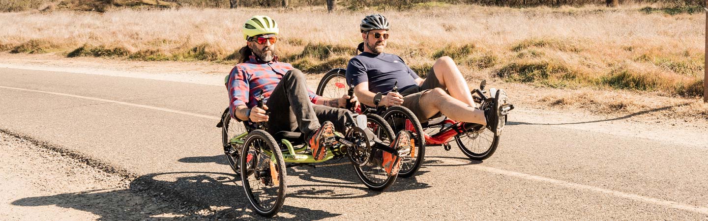 Two men riding recumbent trikes next to each other on a trail