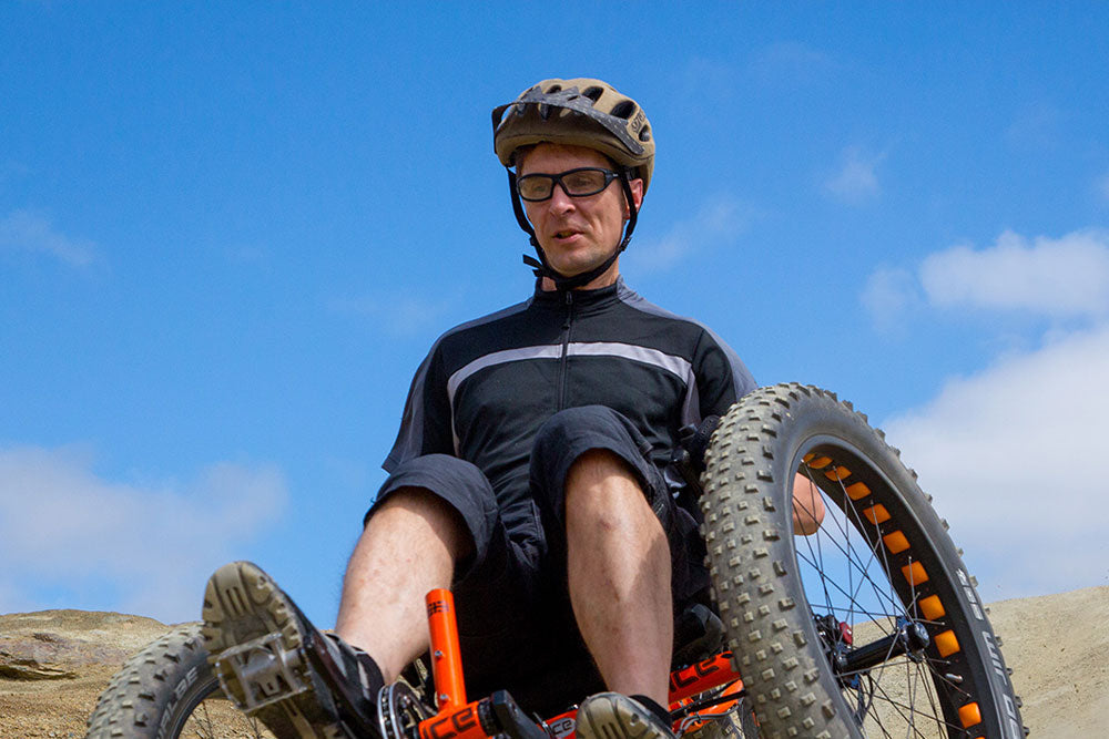 Rider on an orange ICE Full Fat fat-tire recumbent trike climbing a sandy off-road berm under a blue sky