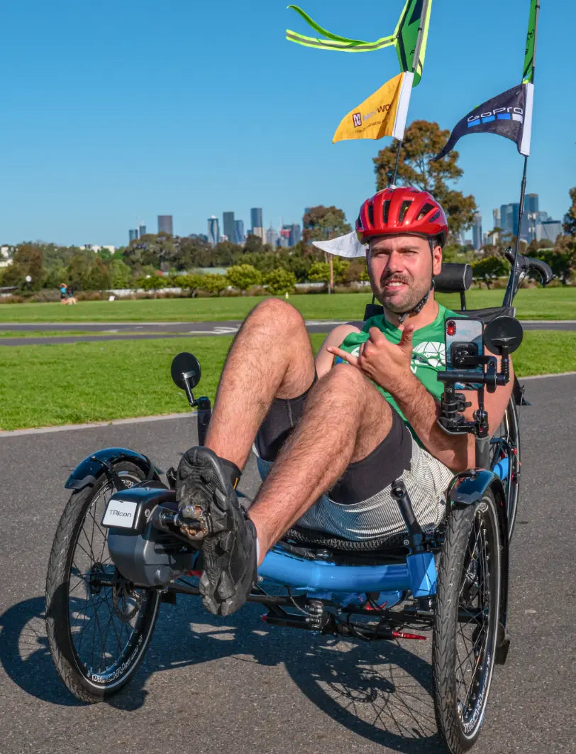 Rider on a blue AZUB TRIcon 26 recumbent trike on a park path with a city skyline behind