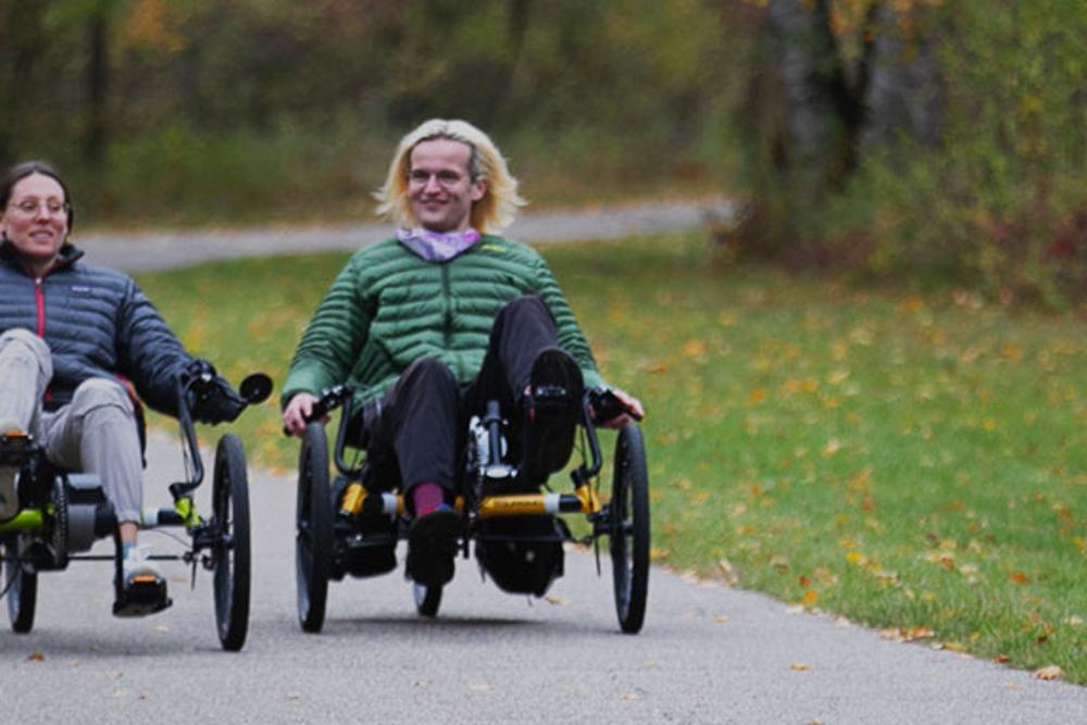 Two riders on TerraTrike E.V.O. electric recumbent trikes on a tree-lined path