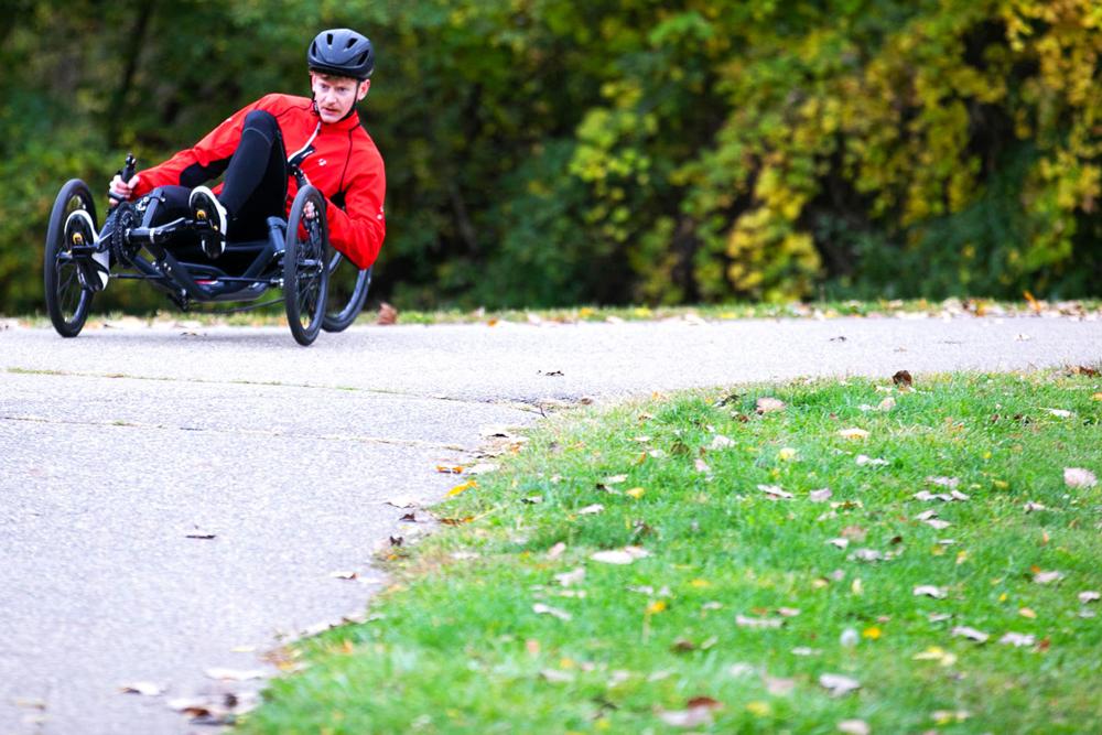 Man in a red jacket riding a TerraTrike Spyder recumbent trike on an autumn leaf-covered path