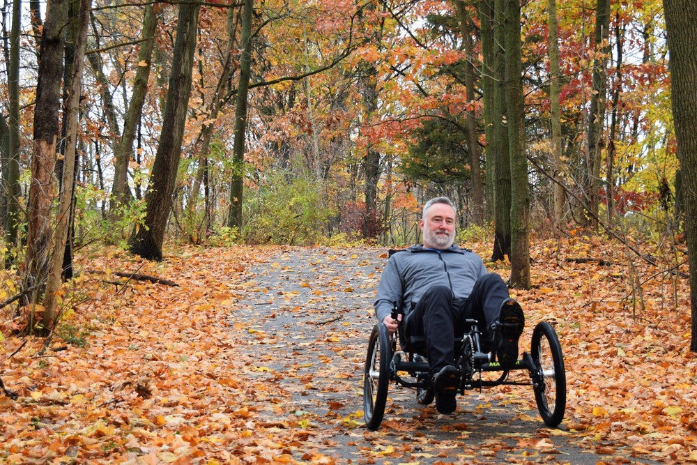 Rider on a GreenSpeed Magnum XL recumbent trike on an autumn leaf-covered trail