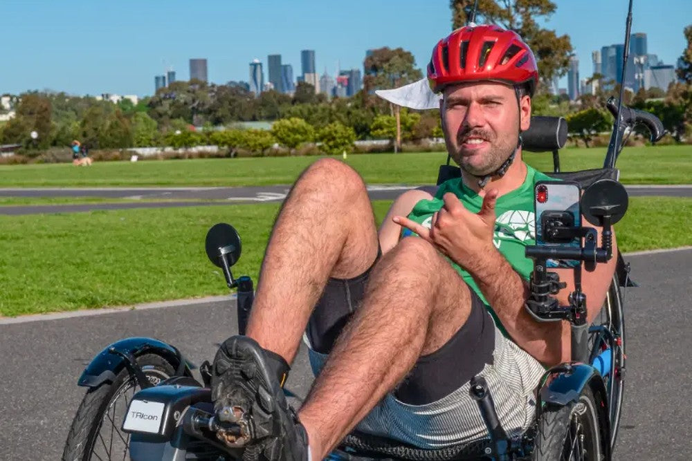 Rider on a blue AZUB TRIcon 26 recumbent trike on a park path with a city skyline behind