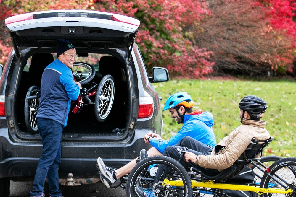 Rider loading a folded TerraTrike Traveler recumbent trike into the back of an SUV while two riders wait on a fall path