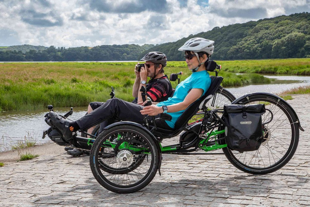 Two riders touring on green ICE Adventure recumbent trikes with panniers beside a lake