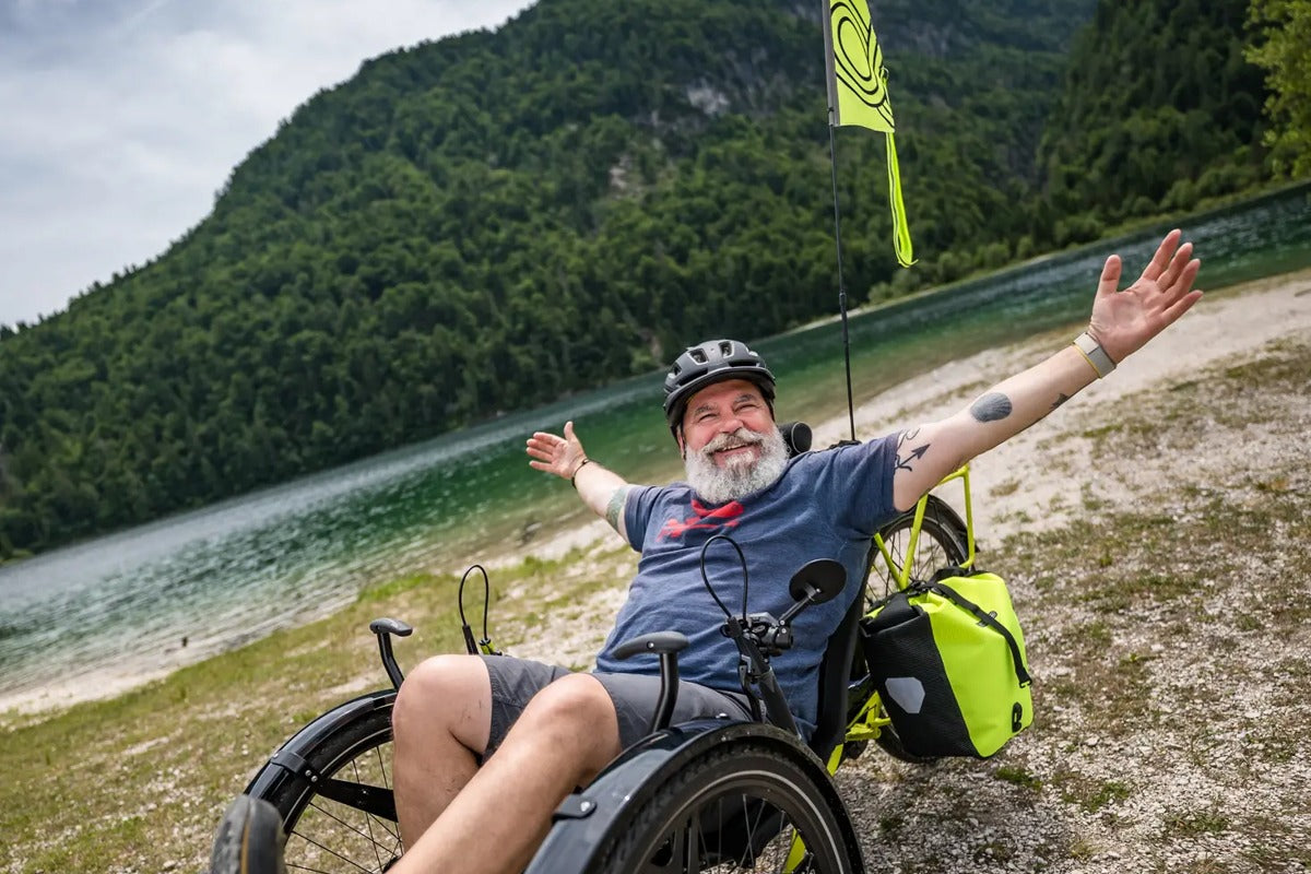 Smiling rider stretching both arms wide on an AZUB Ti-FLY X full-suspension recumbent trike beside a mountain lake